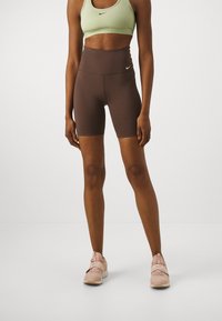 Hnědé cyklistické šortky s vysokým pasem a světle zelená sportovní podprsenka s přiléhavým střihem a logy Nike, doplněné růžovými sportovními botami na jednobarevném pozadí.