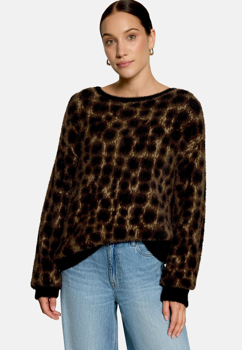 Femme portant un pull duveteux à motif léopard avec des poignets noirs et un jean bleu clair, debout devant un fond uni.