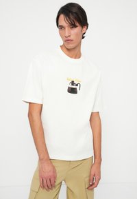White cotton t-shirt featuring a graphic of a black coffee pot with yellow text saying "Café du Choix." Short sleeves and standard fit.