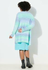 Neulejakku sinivihreässä gradienteissa, pitkät hihat, avonainen etuosa ja pidempi takakaarre, yhdistetty turkoosiin mekkoon ja mustiin nilkkasaappaisiin.