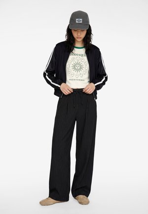 Junge Person trägt eine graue Mütze, schwarze Reißverschlussjacke mit weißen Streifen, cremefarbenes T-Shirt mit Sternzeichenmotiv, schwarze weite Hose mit Nadelstreifen und beige Schuhe.