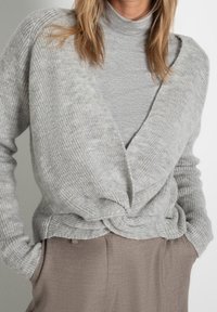 Femme portant un cardigan en maille gris clair enroulé sur un pull à col roulé gris et un pantalon taupe, les mains dans les poches.