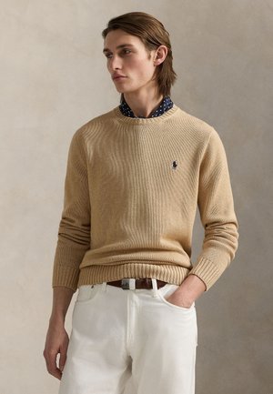 Jeune homme aux cheveux bruns portant un pull beige par-dessus une chemise bleu marine à pois et un pantalon blanc, une main dans la poche.