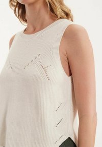 Top sans manches tricoté en crème avec un design texturé, des accents de mailles ouvertes sur les côtés et un col côtelé.