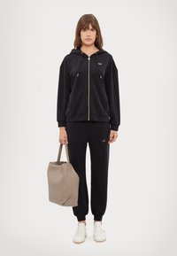 Felpa nera con zip e dettagli dorati, pantaloni joggers neri abbinati e sneakers bianche. Borsa beige con design strutturato.