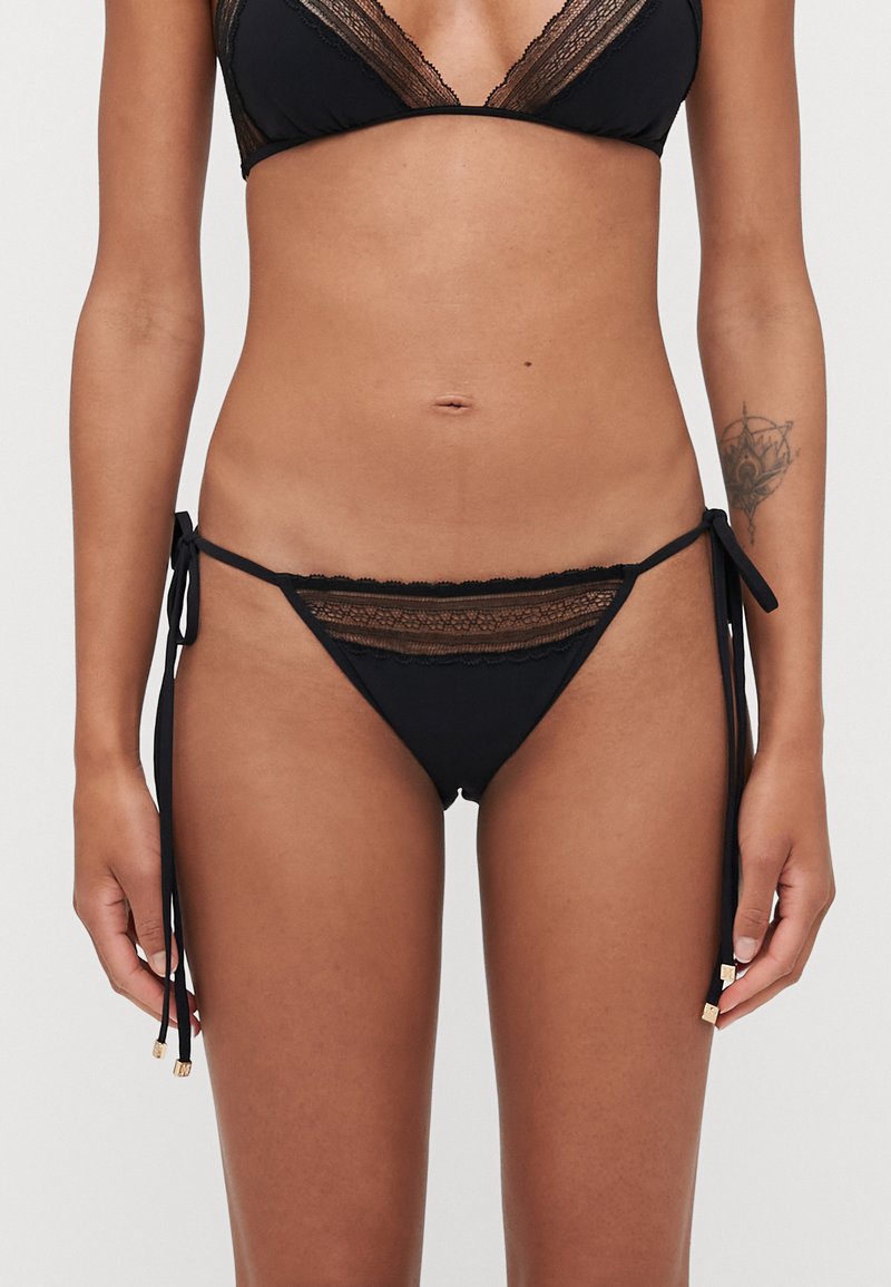 Black bikini bottom with lace accents and tie sides. Features gold hardware on ties and a low-rise design with soft texture.