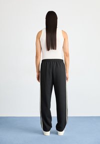 Černé tepláky s bílými bočními pruhy, elastickým pasem a manžetami. Modelka má na sobě přiléhavé bílé tílko, pohled ze zadu.