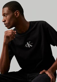 Camiseta de algodón negra con un logo bordado en blanco "CK" en el pecho, mangas cortas y un diseño clásico de cuello redondo.