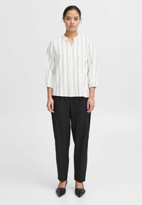 Witte textuurblouse met verticale zwarte strepen, voorzien van een kleine staande kraag en driekwart mouwen, gecombineerd met zwart taps toelopende broeken.