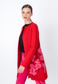 Červený cardigan s kvetinovým dizajnom v ružovej farbe. Vyrobený z mäkkého materiálu, s okrúhlym výstrihom a otvoreným predným dielom. V kombinácii s ružovými nohavicami.