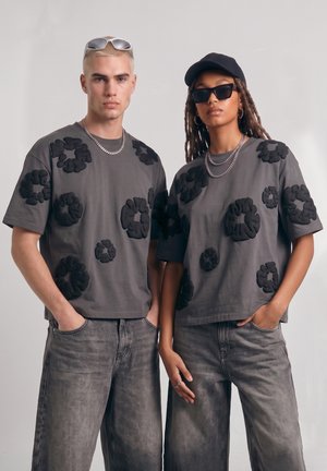 To modeller iført matchende grå mønstrede t-shirts, grå jeans, sølvkæder, den ene med solbriller og kasket, stående med hænderne i lommerne.