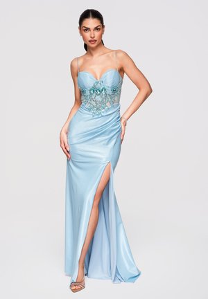 Vrouw in lichtblauwe satijnen jurk met kralenversierd lijfje, dunne bandjes, hoge split tot aan de dij en zilveren sandalen met hoge hakken, poserend tegen een witte achtergrond.