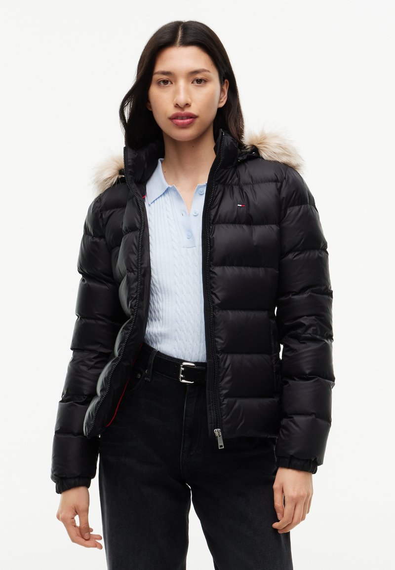 Mujer lleva una chaqueta negra acolchada con capucha de piel sobre una camiseta polo azul claro y pantalones negros, de pie contra un fondo blanco liso.