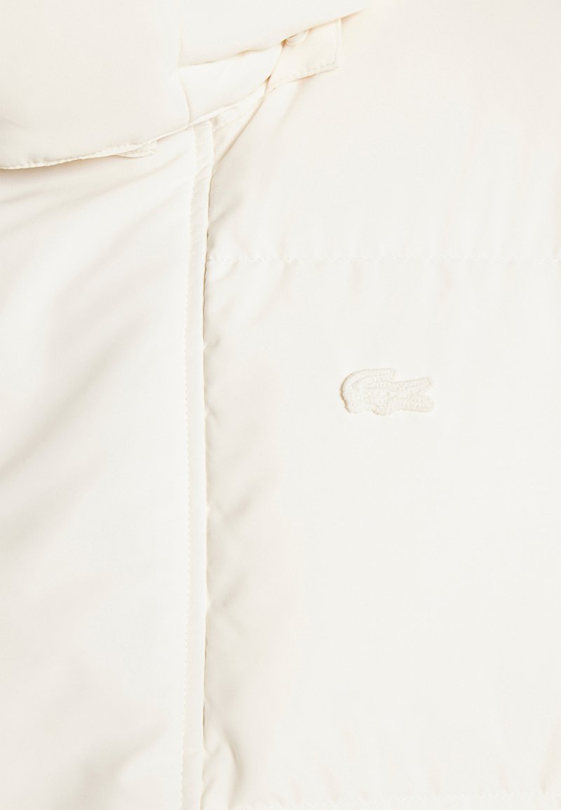 Doudoune blanche avec une texture lisse et brillante, arborant un emblème brodé d'un logo de crocodile sur le devant.