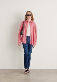 Camisa oversized às riscas vermelhas e brancas, t-shirt branca, jeans azul escuro e sandálias pretas de deslizar. Bolsa simples e óculos de sol.