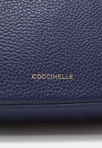 Portafoglio in pelle blu navy con superficie testurizzata, con il logo dorato "Coccinelle" impresso sul davanti. Forma rettangolare compatta.
