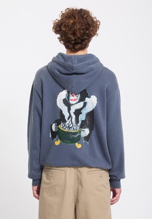 Personne aux cheveux bouclés portant un sweat à capuche gris avec un graphisme d’un squelette en cape noire tenant un chaudron fumant dans le dos.