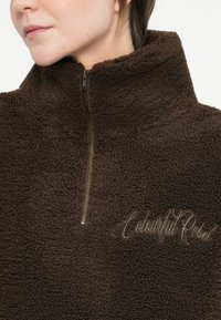Pulover din fleece maro cu guler înalt, închidere cu fermoar și logo-ul brodat "Colourful Rebel" în fir de aur. Textură moale cu un aspect confortabil.