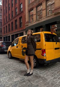 Blazer oversize marrone, camicia bianca con colletto, gonna nera e ballerine nere. Calzini morbidi, strada ciottolata e taxi giallo sullo sfondo.