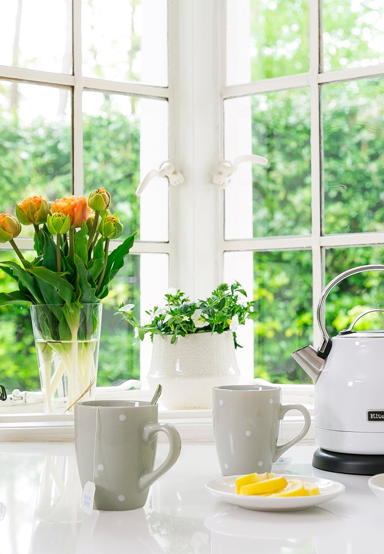 Zwei graue Tassen mit weißen Punkten stehen auf einer weißen Arbeitsplatte neben einem weißen Wasserkocher. Frische Blumen und eine kleine Pflanze sind im Hintergrund sichtbar.
