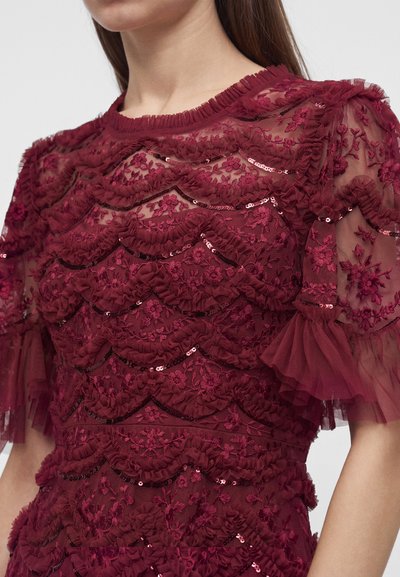 Gros plan sur une robe en dentelle bordeaux pour femme avec broderie florale, détails volantés et sequins sur tissu transparent et manches bouffantes.