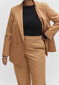 Femme portant un blazer beige et un pantalon taille haute assorti avec un haut noir à manches longues, debout avec une main sur la hanche sur fond blanc.