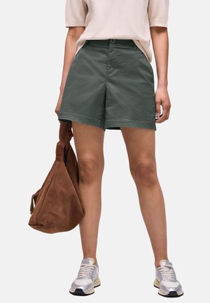 Person iført grønne shorts, beige top, hvide sneakers, der holder en stor brun ruskindstaske ved siden af mod hvid baggrund.