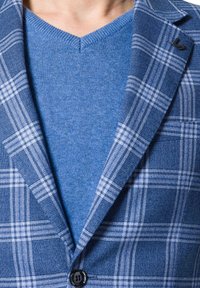 Niebieski marynarka w kratę z wyciętymi klapami i pojedynczym guzikiem, połączona z jednolitym niebieskim swetrem w serek. Teksturowany materiał w różnych odcieniach niebieskiego.