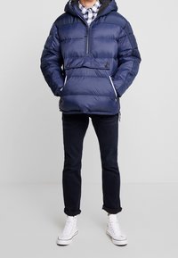 Granatowa kurtka puchowa z zamkiem z przodu, kapturem i dwoma guzikami na piersi. Połączona z ciemnymi jeansami i białymi sneakersami za kostkę.