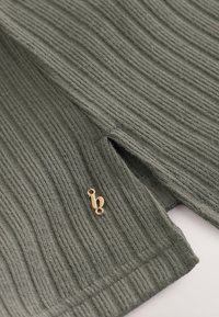 Rippstrickpullover in Grün mit einem goldenen Metallschild, das den Buchstaben "b" trägt. Weicher Stoff mit einem strukturierten Muster über die gesamte Oberfläche.