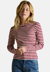 Garais piedurkņu tops ar sarkaniem un baltiem svītrām, izgatavots no kraukšķīga auduma, ar apaļu izgriezumu un pieguļošu dizainu. Apvienots ar parādītajām džinsām.