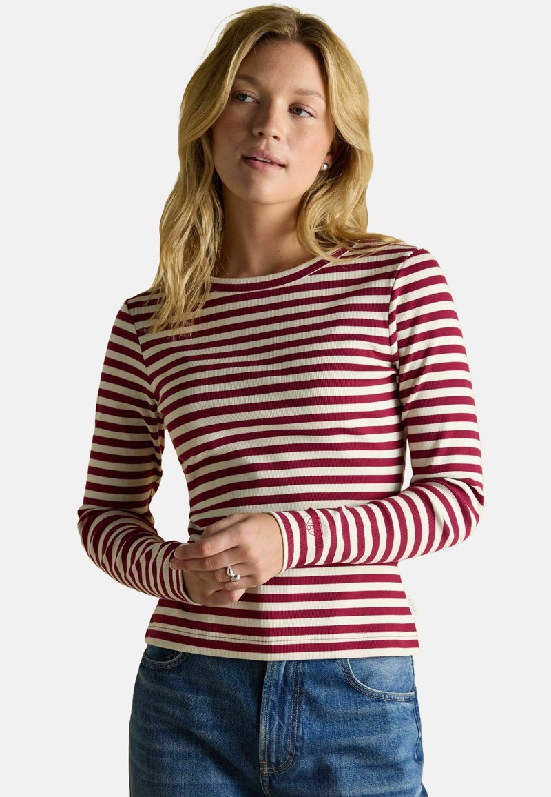 Garais piedurkņu tops ar sarkaniem un baltiem svītrām, izgatavots no kraukšķīga auduma, ar apaļu izgriezumu un pieguļošu dizainu. Apvienots ar parādītajām džinsām.