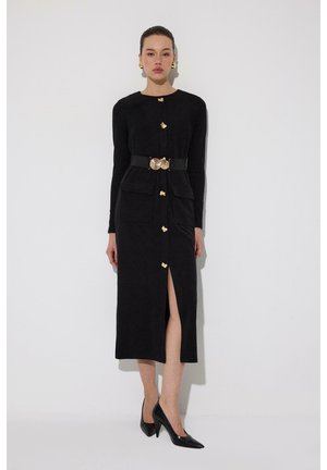 Kvinde iført en lang sort kjole med gyldne knapper, to store lommer foran, et bælte med en cirkulær gylden spænde og sorte højhælede sko.