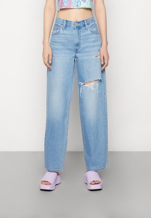 Jean large taille haute bleu clair avec un genou déchiré et un ourlet effiloché. Porté avec des sandales à plateforme lavande.