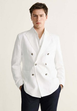 Joven con expresión seria que lleva una blazer blanca cruzada sobre una camisa blanca, manos en los bolsillos, pantalones oscuros.