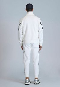 Wei�ßes Sportjacken- und Hosen-Set mit schwarzen Streifen an den Seiten und gesticktem Logo. Das Modell trägt eine dunkle Mütze und silberne Sneakers.