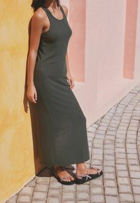 Vrouw in een mouwloze donker groene geribbelde jurk en sandalen, staand tegen een geel-roze muur op een geplaveid pad.