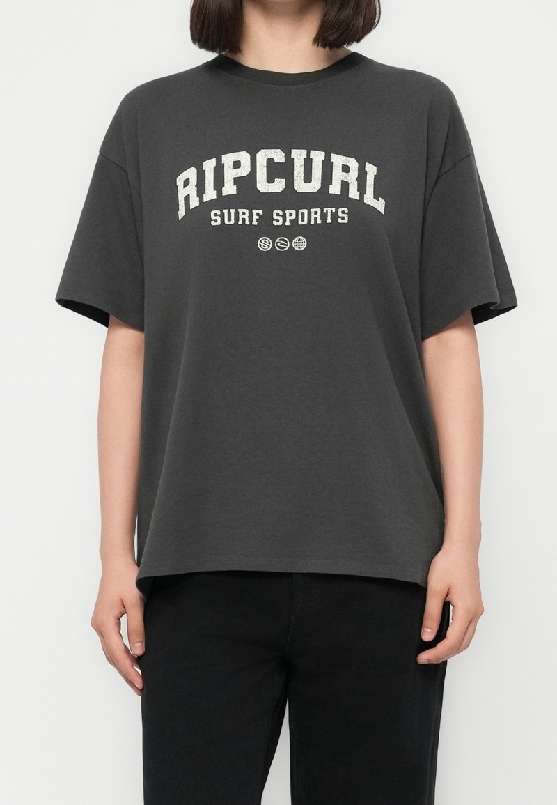 Personne portant un t-shirt ample gris foncé avec le texte « RIP CURL SURF SPORTS » et trois petites icônes circulaires sous le texte.