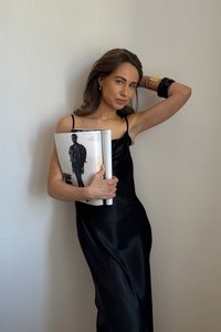 Musta satiininen liukumallinen mekko ohuilla olkaimilla; malli pitää kädessään lehteä, jossa on muotikuva. Sileä pinta ja istuva siluetti.