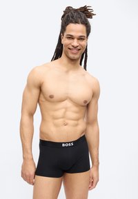 Homme souriant torse nu avec de longues dreadlocks portant un caleçon noir BOSS sur un fond clair uni.