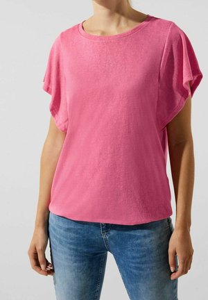 Vrouw draagt een felroze top met korte, losse mouwen en blauwe spijkerbroek, staand tegen een effen lichte achtergrond.