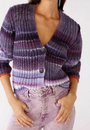 Nainen, joka pukeutuu liila-vaaleanpunaraidalliseen neulottuun neuletakkiin, jossa mustat napit, ja haalistuneen vaaleanpunaisiin korkeavyötäröisiin farkkuihin, kädet taskuissa.