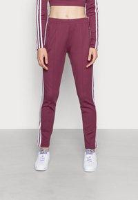 Pantaloni sportivi bordeaux con strisce bianche sui lati, vita elastica e vestibilità slim. Abbinati a sneaker bianche. Tessuto liscio.