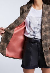 Brązowa marynarka w kratę z różową satynową podszewką, noszona na białej koszulce z nadrukiem i czarnych skórzanych szortach z podwiniętym rantem.