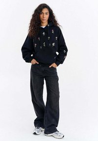 Černá mikina s kapucí s květinovou výšivkou v barvách, přední klokaní kapsa a stahovací šňůrka. Kombinováno se širokými tmavými džíny a teniskami.