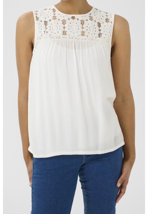Mujer con top blanco sin mangas con canesú de encaje floral y vaqueros azules, de pie con los brazos relajados a los lados contra un fondo liso.