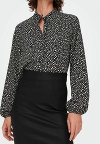 Zwarte blouse met witte abstracte patronen, een knoopsluiting en een kraag. Lange, opgeblazen mouwen gecombineerd met een zwarte, getailleerde rok.