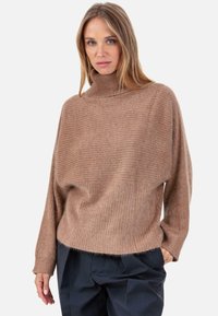 Pull à col roulé côtelé couleur brun clair, coupe ample, avec manches longues et texture douce. Porté sur un pantalon foncé.