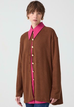 Cardigan marrón con relieve y una camisa de color rosa con cuello debajo, que presenta botones dorados y una silueta oversized. Textura suave y corte relajado.