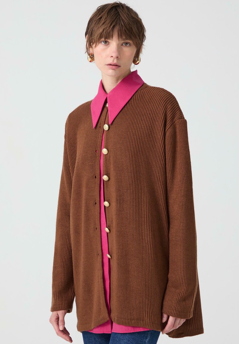 Brown ribbed cardigan with a pink collared shirt underneath, featuring gold buttons and an oversized silhouette. Soft texture and relaxed fit.